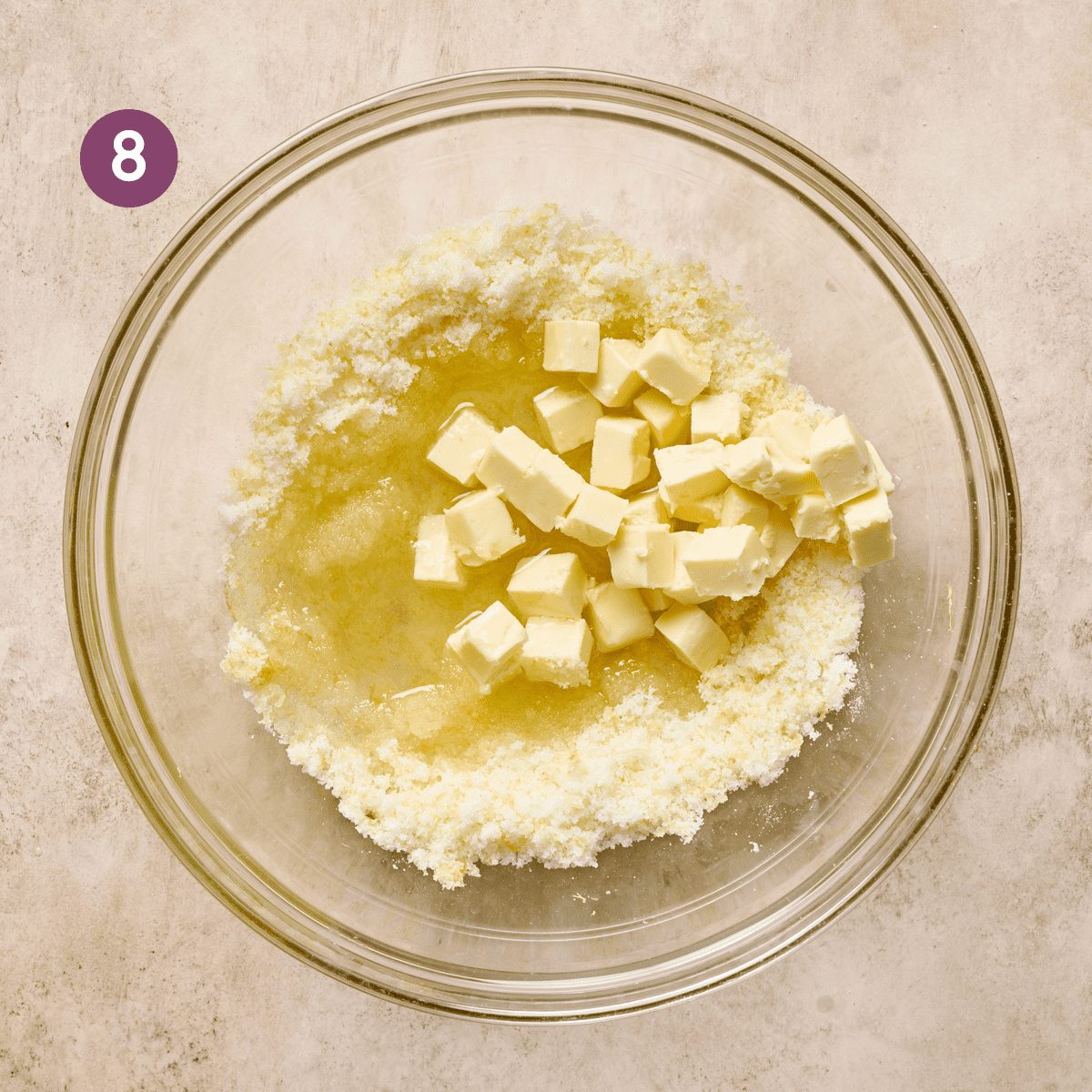 
cubed vegan butter and oil sitting on top of sugar in a large glass bowl on a light pink surface. 